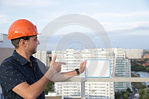 Builder in a protective helmet with a tablet for writing in his hand is standing on the roof of a building overlooking the city