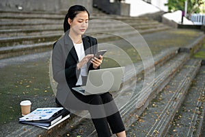 A professional Asian businesswoman is responding to an urgent message while sitting on the stairs