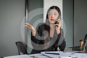 A professional Asian businesswoman is talking on the phone with her client while sitting at her desk