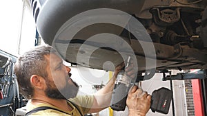 Professional auto mechanic tightens bolts with special electric tool at workshop. Man working underneath a lifting