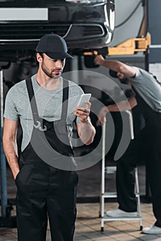 professional auto mechanic using smartphone while colleague working