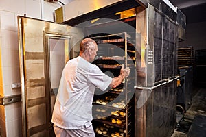 Professional baker in uniform inserts cart with decks for baking raw dough to make bread in an industrial oven in a bakery