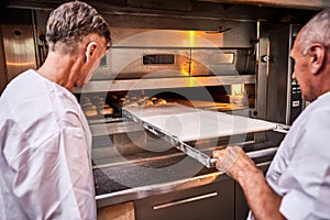 Professional baker in uniform inserts cart with decks for baking raw dough to make bread in an industrial oven in a bakery