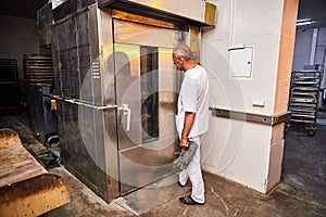 Professional baker in uniform inserts cart with decks for baking raw dough to make bread in an industrial oven in a bakery