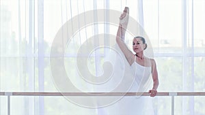 Professional ballerina in white tutu is stretching her legs near the barre stand