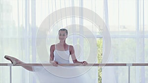 Professional ballerina in white tutu is stretching her legs near the barre stand