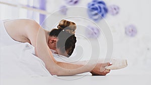 Professional ballerina in white tutu is stretching sitting on the floor.