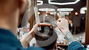 Professional barber with scissors and hair comb in hands making haircut for his client in barber shop. Cropped photo