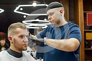 Professional barber serving young handsome man in the modern barbershop, working with hair clipper. Focus on a