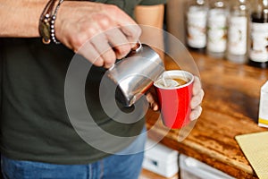 Professional barista man preparing coffee in a modern coffee shop. Male hands holding a metal cup and a paper red cup. Closeup
