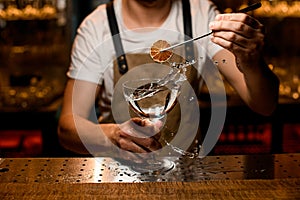 Professional bartender adding to an alcoholic cocktail a dried lemon slice with tweezers