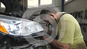 Professional bearded repairman fixing car with open hood at workshop. Auto mechanic in uniform working with vehicle at