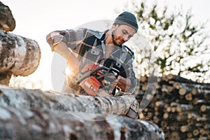 Professional bearded strong lumberman wearing plaid shirt sawing tree with chainsaw for work