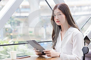 Professional beautiful young Asian business woman is looking at camera in her hands tablet or notepad while she is sitting at