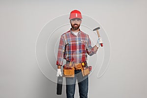 Professional builder in hard hat with hammer and tool belt on light background