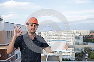Builder in a protective helmet with a tablet for writing in his hand is standing on the roof of a building overlooking the city