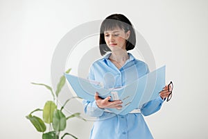 Professional businesswoman in formal blue shirt checks paper documents in folder stands on white wall background with