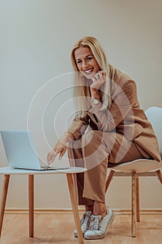 A professional businesswoman sits on a chair, surrounded by a serene beige background, diligently working on her laptop