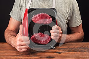 Professional butcher in grey t shirt and classic red and white stripe apron holding plastic prepacked tray with two premium fillet