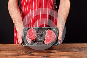 Professional butcher in grey t shirt and classic red and white stripe apron holding plastic prepacked tray with two premium fillet