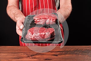 Professional butcher in grey t shirt and classic red and white stripe apron holding plastic prepacked tray with two premium fillet