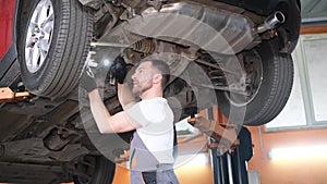 A professional car mechanic works under the king of a hoisting machine in a car repair shop.