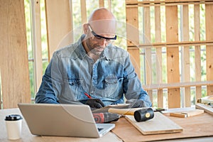 Professional carpenter man wearing glove and goggle writing some idea into clipboard in the modern wood workplace. Many equipment