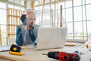 Professional carpenter wear glove sitting on the chair and hold pencil and looking at laptop. Working in the modern wood workplace
