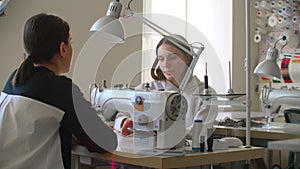 Professional Caucasian dressmaker and young seamstress choosing fabric and talking while colleague using sewing machine