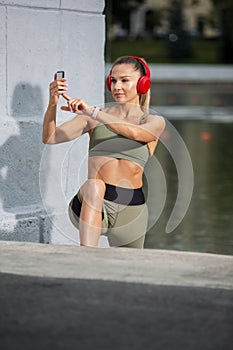 Professional Caucasian Female Athlet In Running Outfit During Her Stretching Exercises Outdoor. Listening Music in Headphones