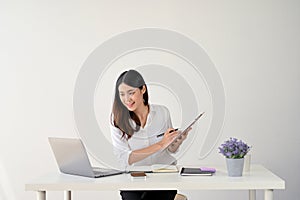 A businesswoman is taking notes on paper on a clipboard while looking at her laptop screen