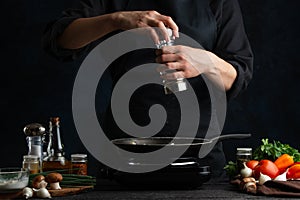 The professional chef in black uniform pours spices in pan with cooking meal on the stove. Backstage of preparing grilled meat for