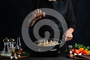 The professional chef in black uniform pours spices in pan with meat. Backstage of preparing grilled meat for dinner on dark blue