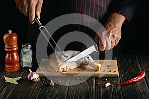 Professional chef cuts a fresh raw chicken drumstick on a dark background. Nearby on the kitchen table are ingredients for cooking