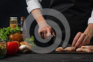 Professional chef cuts with knife chicken fillet on black chopped board with variety of ingredients background. Backstage of