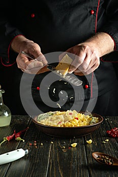 A professional chef grates hard cheese onto a plate with pasta using a hand grater. Working environment in a hotel kitchen with