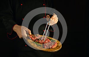 Professional chef holds a fork in his hand and a cutting board with beef steaks before barbecue. Dark space for restaurant recipe