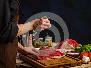 Professional chef prepares juicy steaks from fresh meat on a wooden cutting board, dark background. Sprinkle the pieces of meat