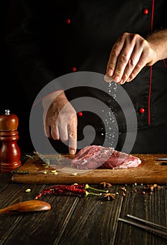 Professional chef sprinkles salt into meat steak for cooking for barbecue lunch. Work environment on the kitchen table in a