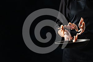 The professional chef toss up raw chicken legs above the frying pan on black background. Backstage of cooking grilled meat for