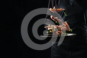 The professional chef toss up raw chicken legs with asparagus above the frying pan on black background. Backstage of cooking