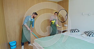 Professional cleaners in uniform washing floor and wiping dust from the furniture in the hotel room or apartment bedroom