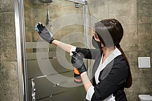 Professional Cleaning service company employee in rubber gloves with spray bottle and blue rag cleaning a shower in bathroom