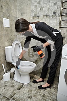 Professional Cleaning service company employee in rubber gloves with spray bottle and blue rag cleaning a toilet bowl in bathroom