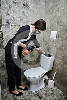 Professional Cleaning service company employee in rubber gloves with spray bottle and blue rag cleaning a toilet bowl in bathroom