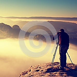 Professional on cliff. Nature photographer takes photos with mirror camera on rock. Dreamy fogy landscape, spring orange pink mist