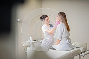 Professional cosmetician examining face skin of girl in clinic of esthetic cosmetology