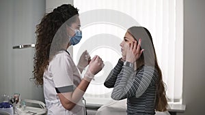 Professional cosmetologist in white lab costume and mask at working place meets the client - young girl with long hair