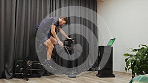 Professional cyclist cycling out of saddle and drinking energetic drink or water. Man pedaling at home on stationary smart bicycle