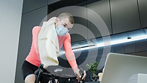 Professional cyclist in protective medical face mask is pedaling and training on smart stationary cycling trainer while watching o